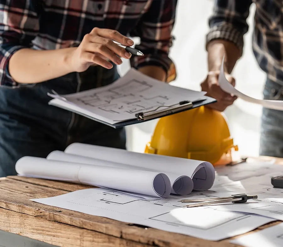 Two people reviewing floor plans on a clipboard beside a hard hat and rolled blueprints